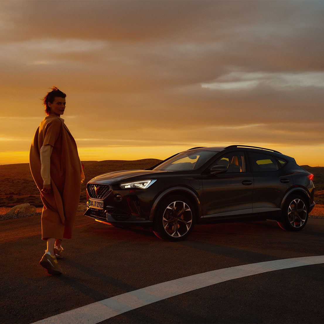 Eine Frau steht vor einem CUPRA Formentor und schaut in die Kamera. Im Hintergrund ist der gelbliche Himmel und wüstenartige Berge zu erkennen.
