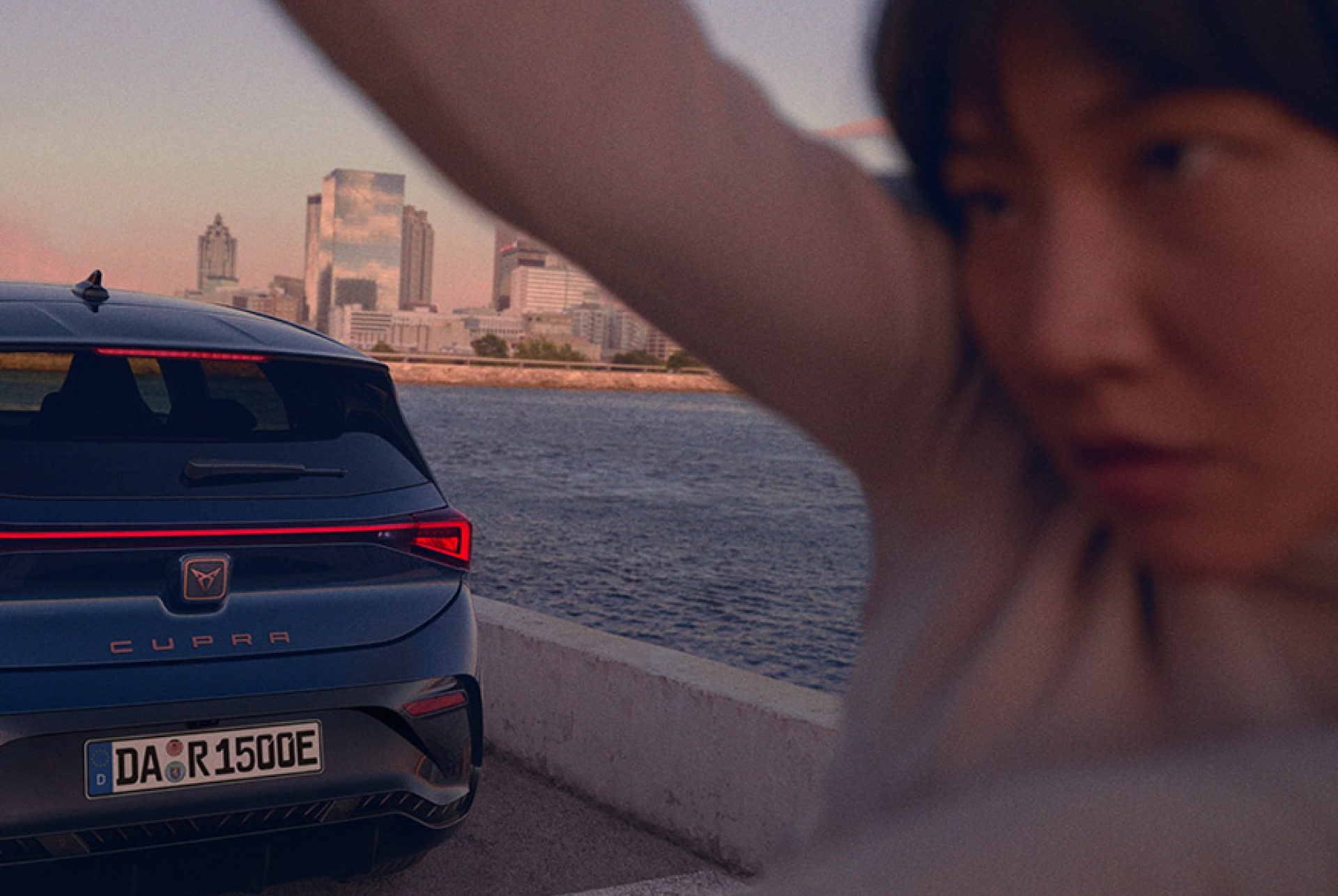 Ein blauer CUPRA. Born steht an einer Uferpromenade. Man erkennt die Hälfte der Rückseite des Autos und vor ihm eine Frau. Im Hintergrund ist das Meer, Strand und Hochhäuser zu erkennen.