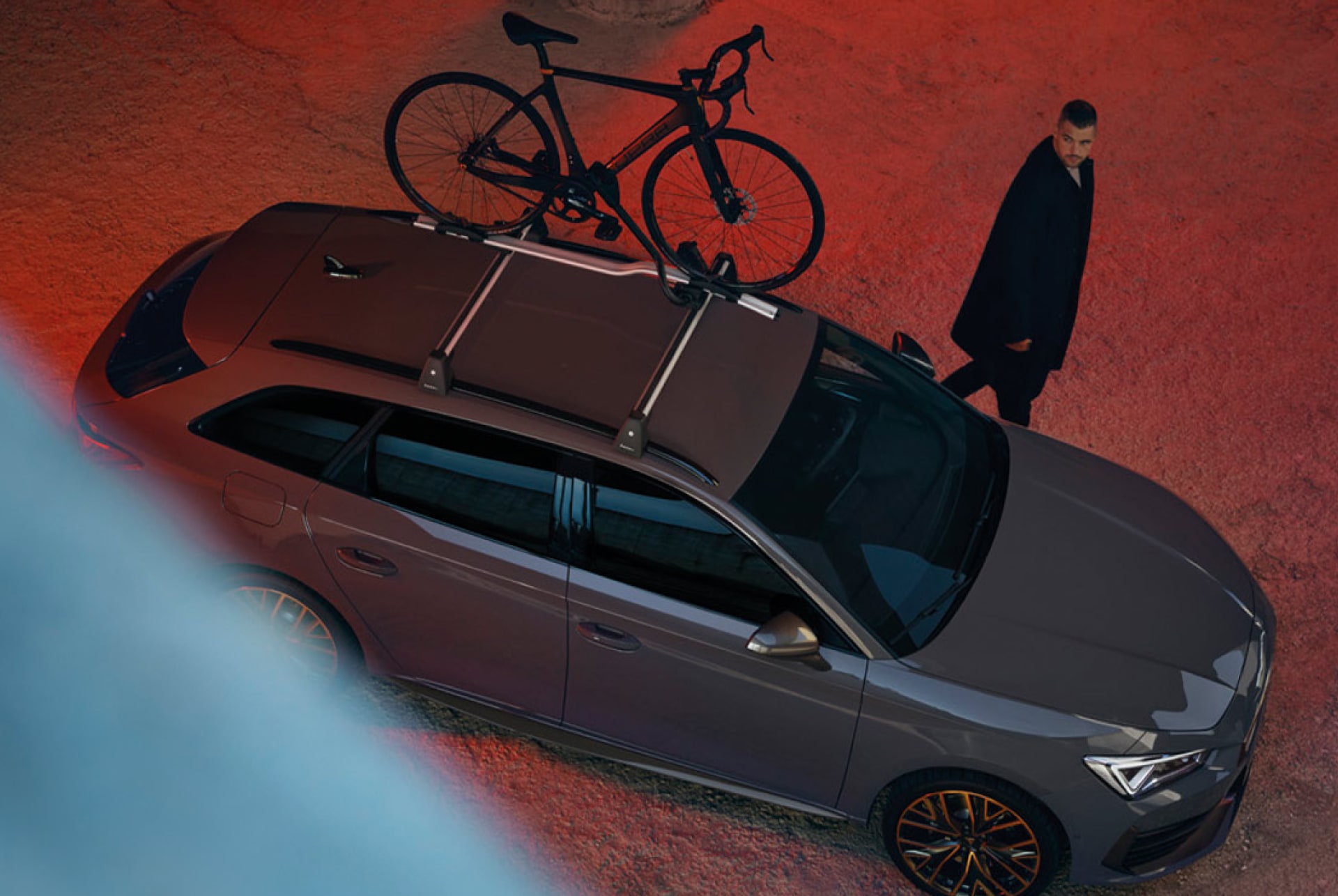 Ein CUPRA Leon Sportstourer in Grau, Vogelperspektive. Auf dem Dach des Fahrzeugs ist ein Fahrrad befestigt, während hinter dem Auto ein Mann in Richtung der Vorderseite des Autos läuft. 