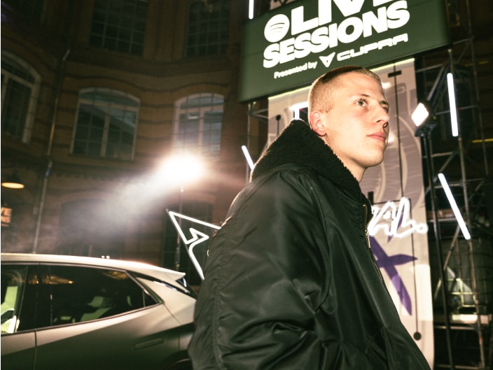  Ein Mann in schwarzer Bomberjacke steht vor dem beleuchteten Schild der „Live Sessions presented by CUPRA“, daneben ist ein CUPRA Fahrzeug zu sehen.