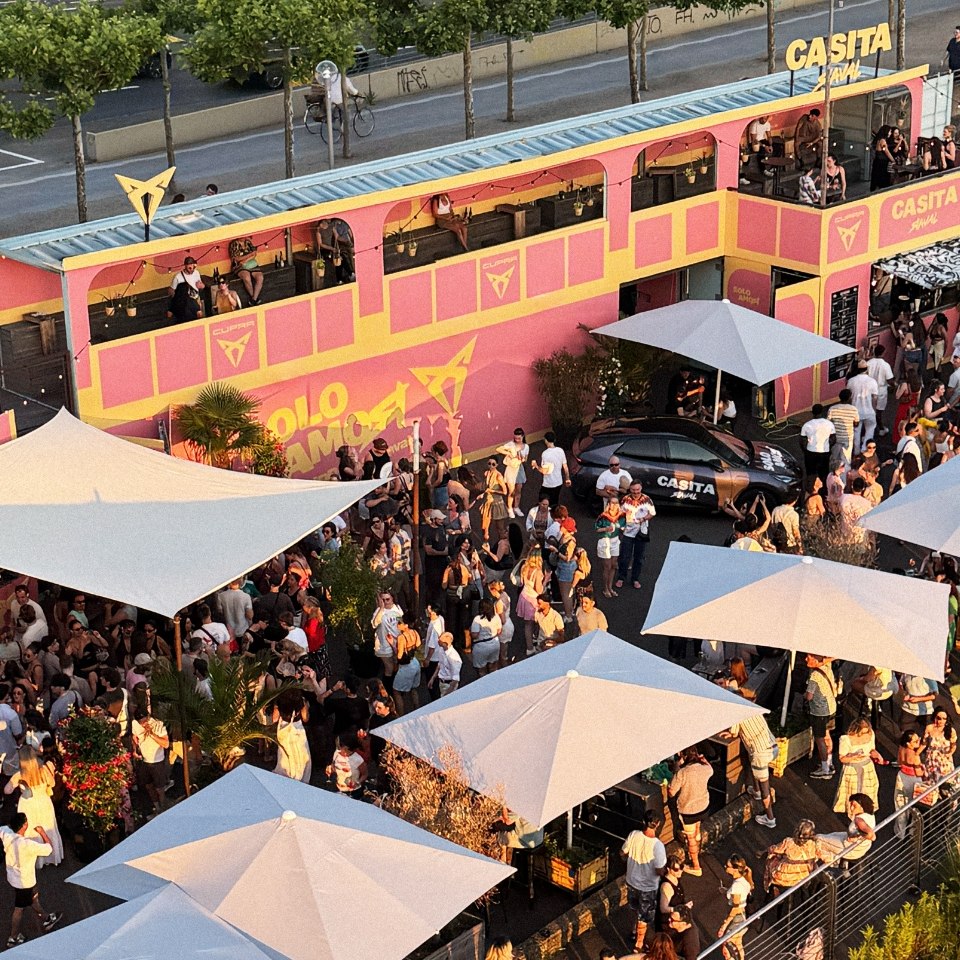 Luftaufnahme der CUPRA Casita Raval in Düsseldorf: pink-gelber Doppeldecker-Pavillon, weiße Sonnenschirme und feiernde Menschenmenge am Rheinufer.