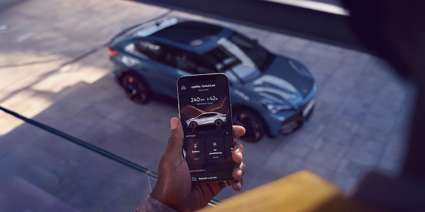 Ein CUPRA Tavascan steht auf einem Parkdeck. Im Vordergrund hält eine Person ihr Smartphone, mit der geöffneten My CUPRA App.  