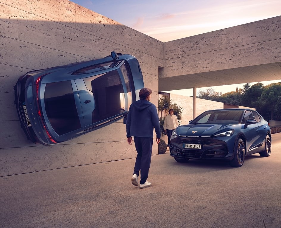 Eine surreale architektonische Szene, in der ein blauer CUPRA senkrecht an einer Betonwand befestigt ist, neben einem anderen blauen Auto, das ganz normal in einer Einfahrt geparkt ist, während eine Person auf den Eingang zugeht.
