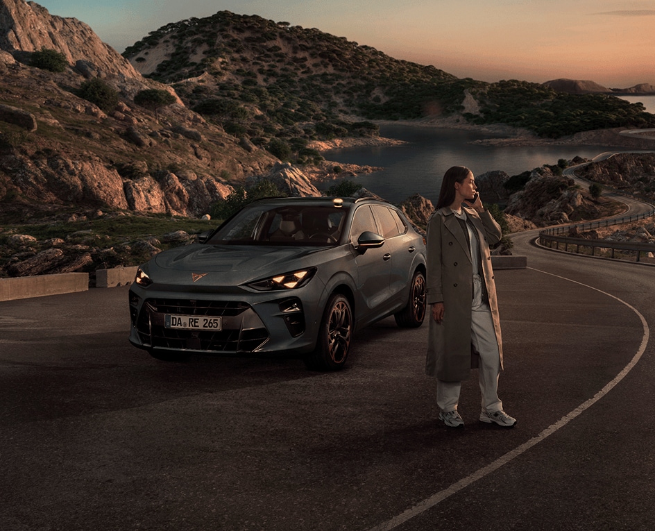 Eine Weitwinkelaufnahme einer Landschaft in der Abenddämmerung, die eine Frau in einem Trenchcoat zeigt, die auf einer kurvenreichen Küstenstraße neben einem CUPRA Terramar steht, im Hintergrund sind felsige Hügel und ein ruhiges Meer zu sehen.