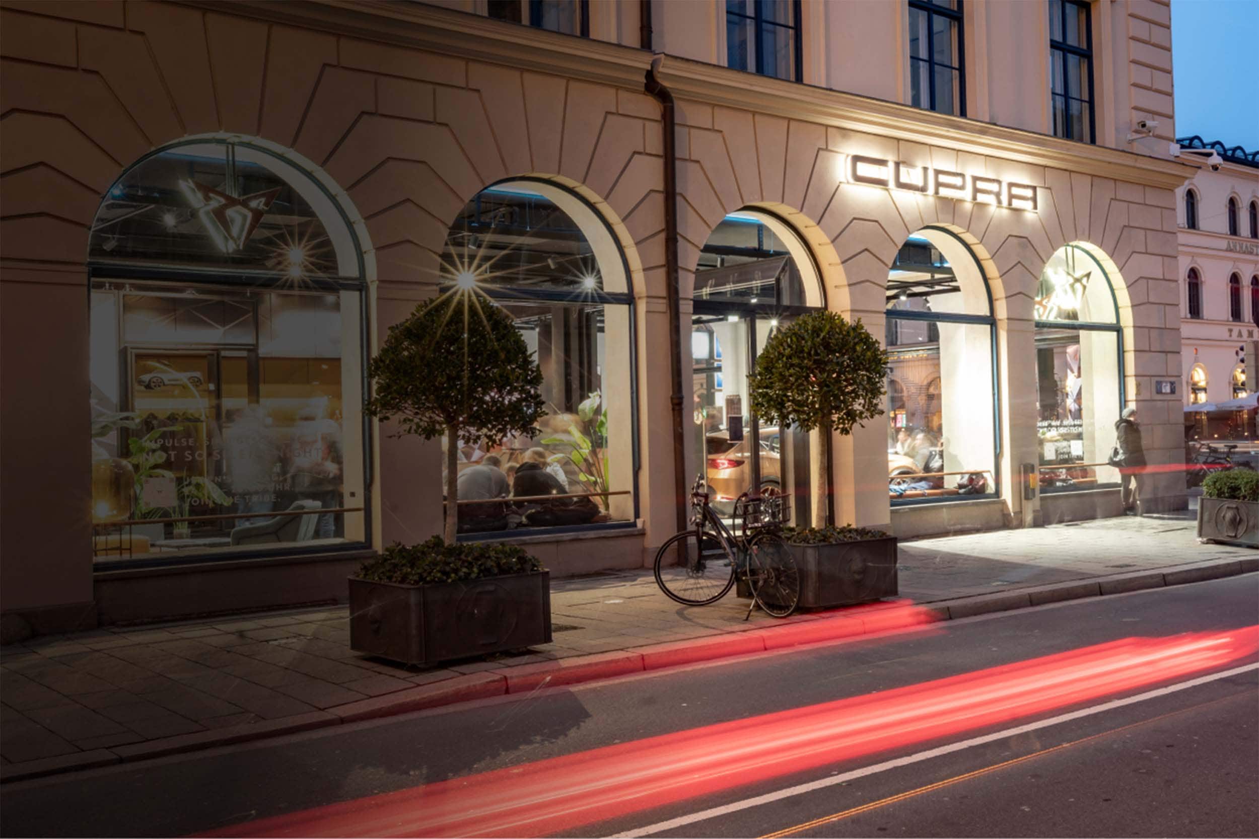Außenansicht der CUPRA City Garage München bei Abenddämmerung mit beleuchtetem Logo, großen Schaufenstern und vorbeiziehenden Lichtspuren auf der Straße.