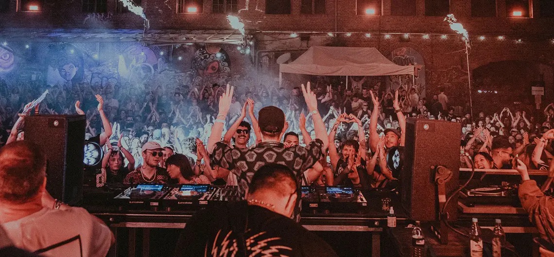 DJ am Mischpult vor einer großen Crowd mit erhobenen Händen in einer Indoor Location.