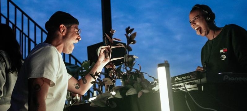 Zwei DJs lachen sich an, rechts ein DJ mit Kopfhörern am Pioneer Mischpult, aufgenommen draußen bei Abendhimmel mit Studio Licht im Vordergrund.