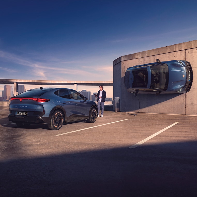 Zwei CUPRA Tavascan Elektrofahrzeuge stehen auf einem Parkdeck, eines wird über ein Ladekabel geladen, das andere ist vertikal an einer Betonwand positioniert.