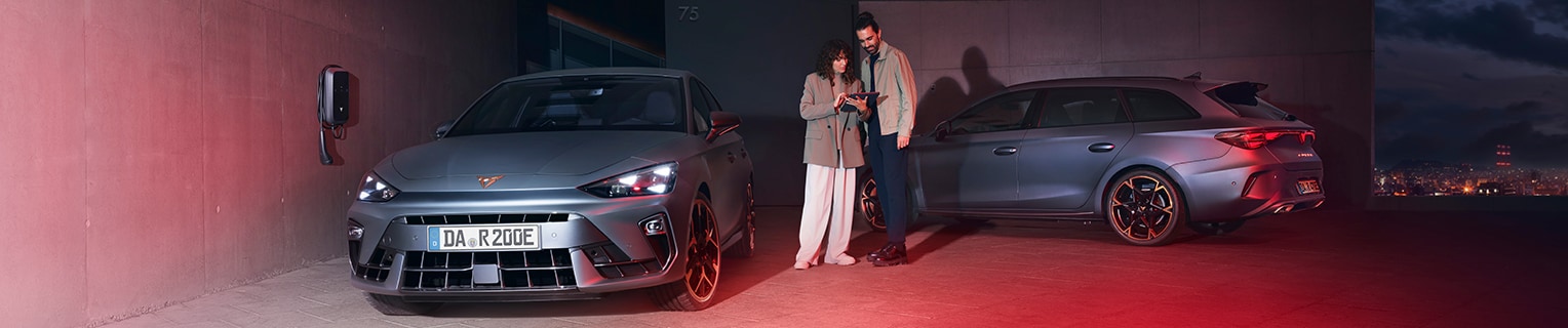 Ein CUPRA Leon und ein CUPRA Leon Sportstourer Plug-in-Hybrid in Mattgrau stehen auf einem beleuchteten Parkdeck mit E-Ladestation. Eine Verkäuferin zeigt einem Interessenten Details auf einem Tablet. Im Hintergrund erstreckt sich eine nächtliche Stadtlandschaft.