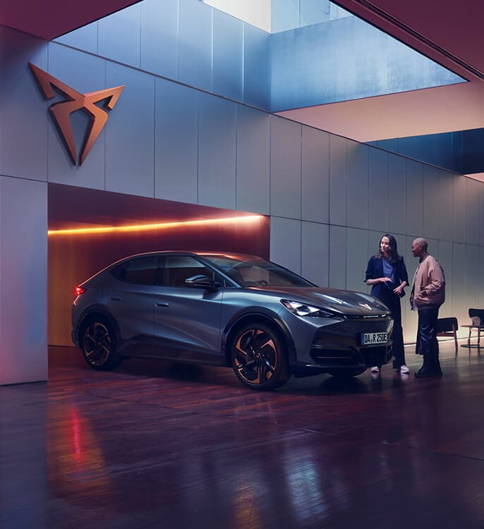 Ein CUPRA Tavascan steht in einem modernen Showroom mit stilvoller Beleuchtung und großem CUPRA-Logo an der Wand. Zwei Personen stehen neben dem vollelektrischen SUV-Coupé und unterhalten sich – es handelt sich um eine Beratungssituation im hochwertigen, urbanen Ambiente.