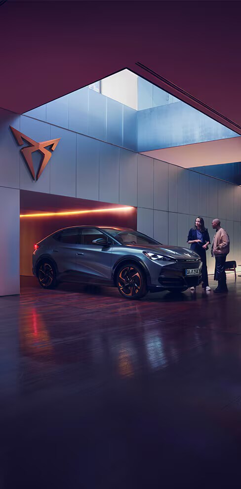 Ein CUPRA Tavascan steht in einem modernen Showroom mit stilvoller Beleuchtung und großem CUPRA-Logo an der Wand. Zwei Personen stehen neben dem vollelektrischen SUV-Coupé und unterhalten sich – es handelt sich um eine Beratungssituation im hochwertigen, urbanen Ambiente.