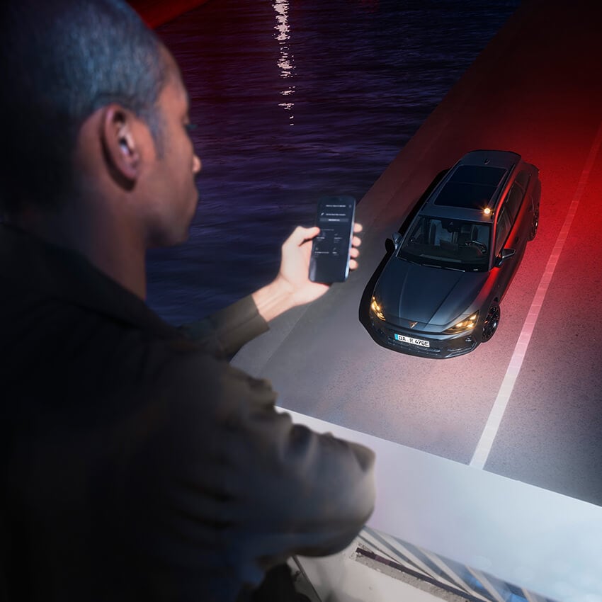 Ein Mann steuert per Smartphone einen CUPRA Leon Sportstourer Plug-in-Hybrid, der am Straßenrand neben einem Wasserkanal parkt. Die Szene bei Nacht zeigt das Fahrzeug von oben mit eingeschaltetem Licht – betont werden Konnektivität, Technologie und modernes Design.