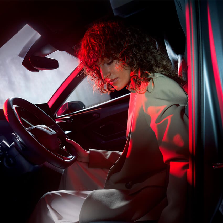 Eine Frau sitzt im Innenraum eines CUPRA Tavascan, elegant gekleidet und von rotem Licht inszeniert. Sie hält das Lenkrad und blickt nach unten – die Szene betont das futuristische Interieur, Design und Fahrerlebnis des vollelektrischen CUPRAs.