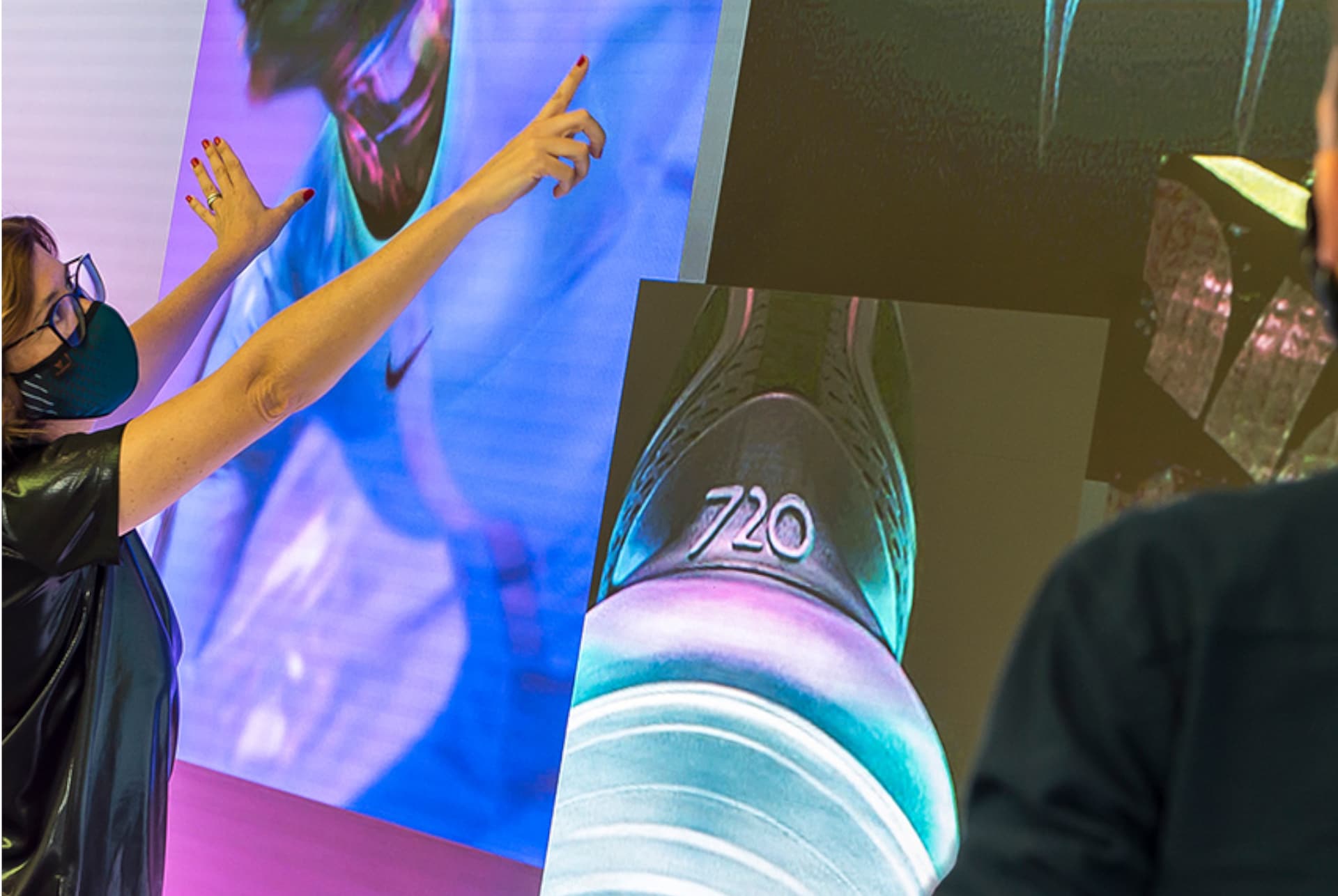 Frau mit Maske deutet auf eine große digitale Leinwand mit farbigen Bildern, darunter eine Nahaufnahme eines Sneakers mit der Zahl 720.