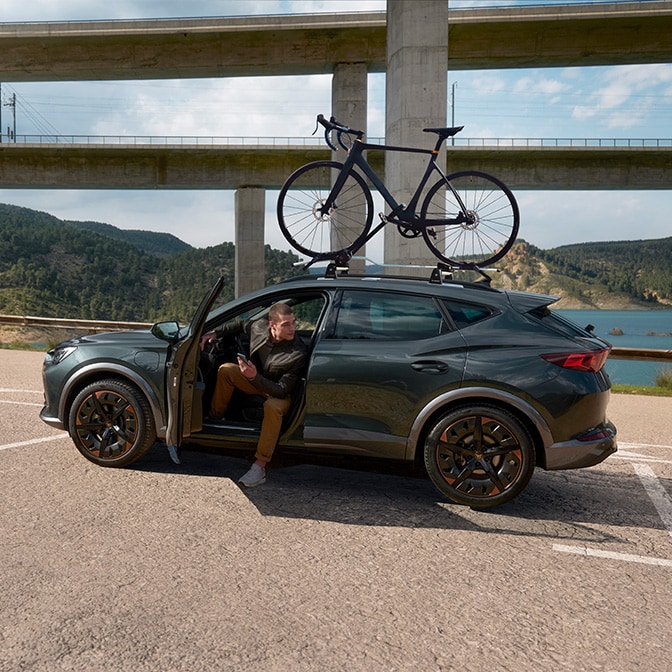 CUPRA Formentor en color Gris Magnetic aparcado cerca de un embalse bajo un puente elevado, con una bicicleta de carretera montada en el techo y una persona saliendo del vehículo en un entorno natural y montañoso.