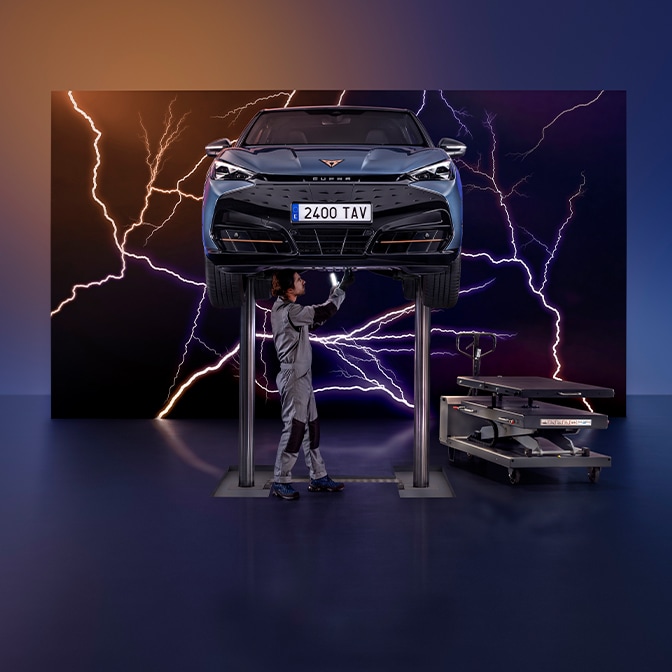 CUPRA Tavascan en el color Tavascan Blue elevado en un taller futurista mientras un mecánico trabaja en la parte inferior, con relámpagos de fondo y ambientación tecnológica.