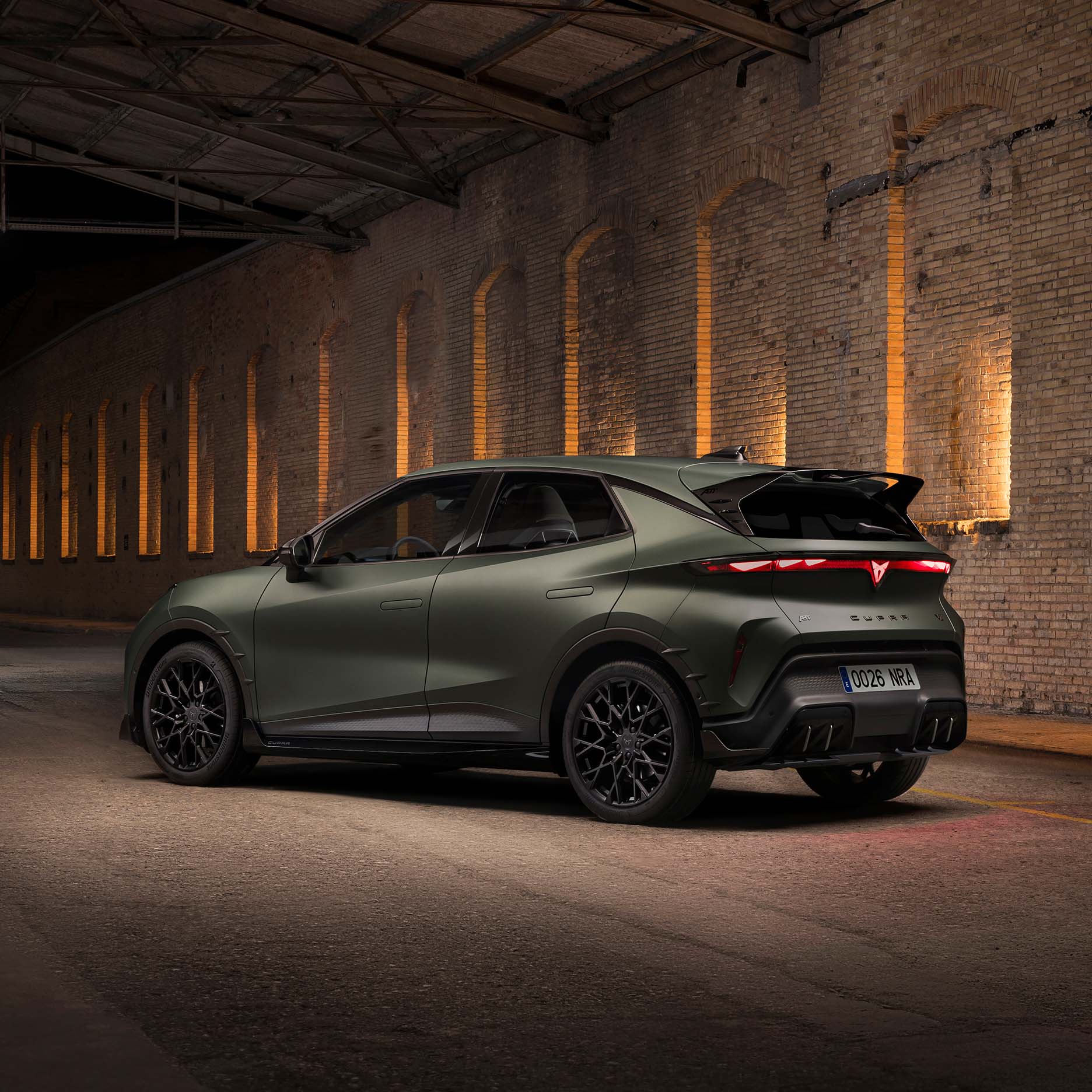 Vue trois-quarts arrière d'une CUPRA Raval Custom Pack stationnée de nuit dans un bâtiment industriel en briques, mettant en valeur son becquet arrière et la signature lumineuse LED rouge.