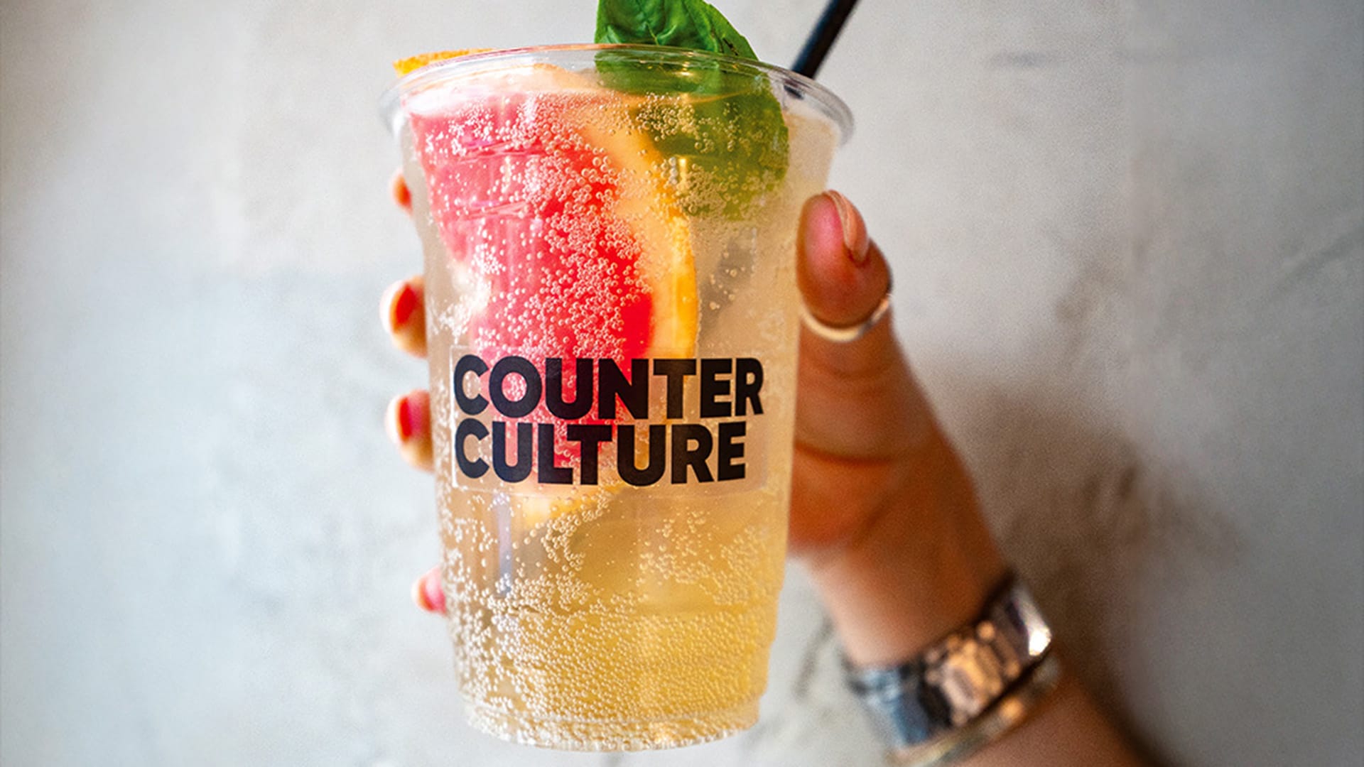 A hand holding a plastic cup of fizzy yellow kombucha with the Counter Culture logo