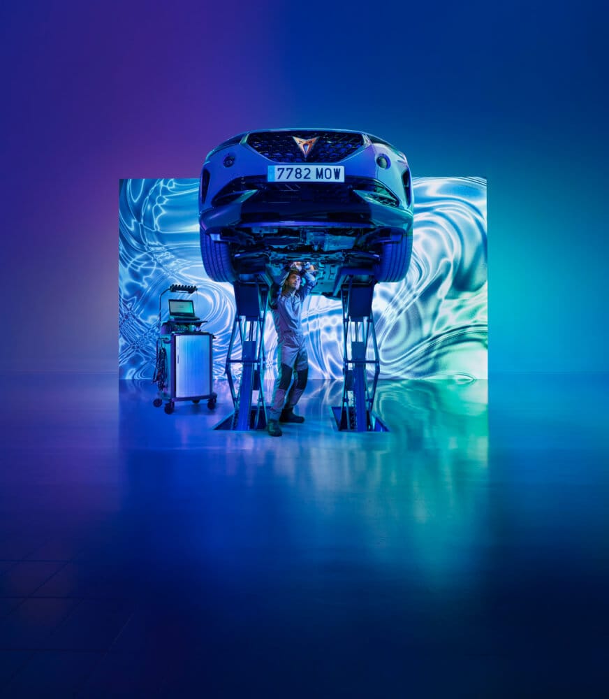 A mechanic in a futuristic, illuminated workshop examines a car on hydraulic lifts. The background displays swirling blue and purple patterns, adding a modern, tech-savvy atmosphere.