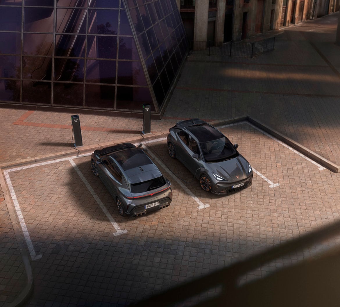 Two CUPRA Raval electric cars charging at kerbside charge points beside a glass building, viewed from above.