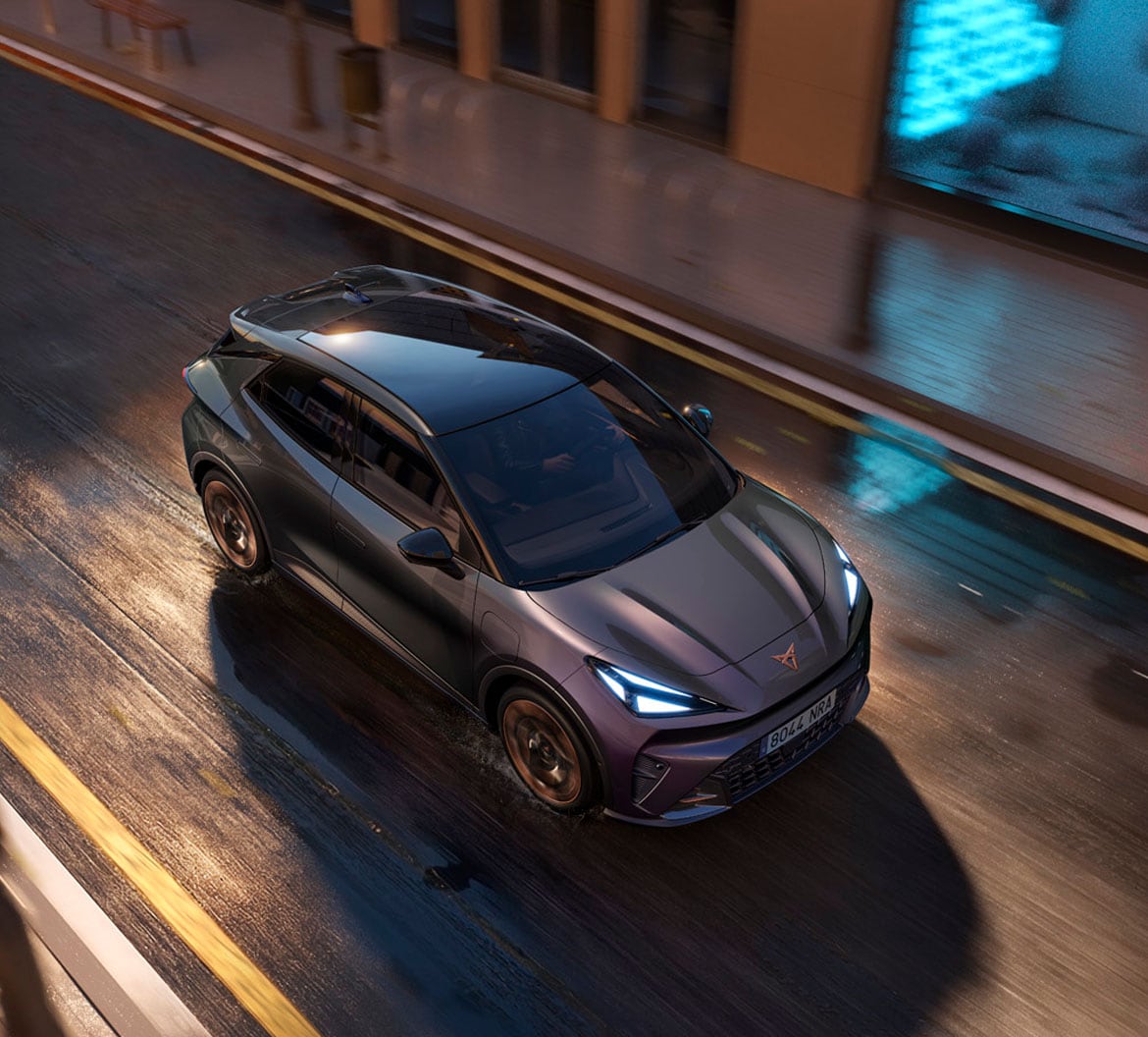 CUPRA Raval electric car driving through a wet city street at dusk, seen from a high angle.