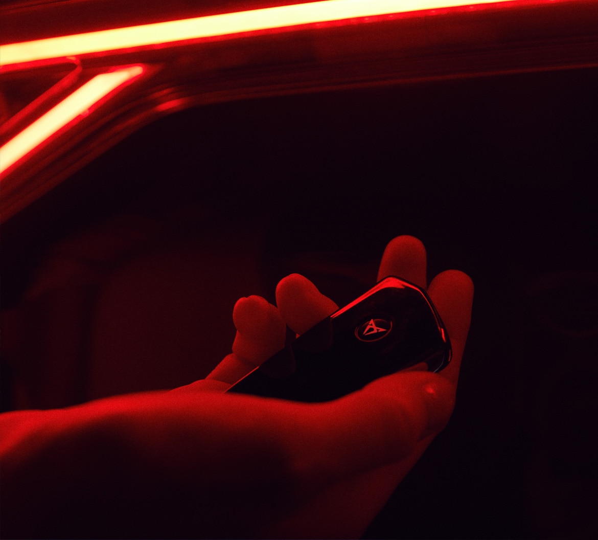A hand holding a CUPRA key fob with the copper triangular logo, lit by dramatic red ambient lighting inside a car.