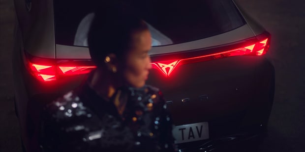 CUPRA Tavascan rear view with illuminated triangular tail lights and a person in a sequined dress at night.