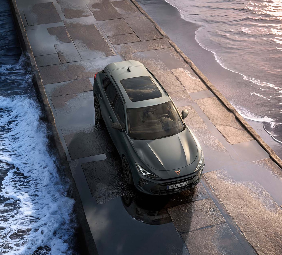 Aerial view of a grey CUPRA Terramar parked on a stone causeway, with waves breaking against both sides at sunset.