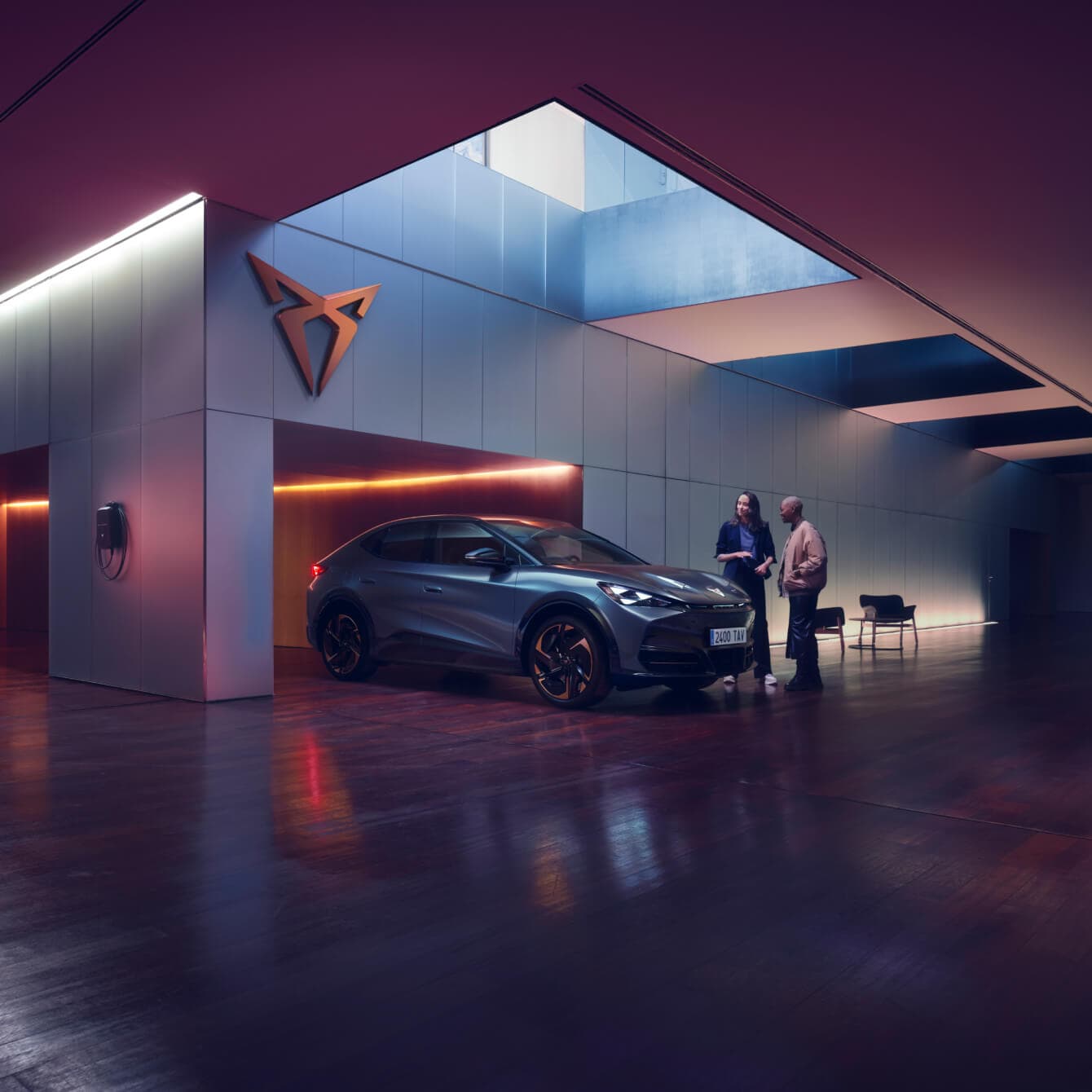 Two people are standing next to a blue CUPRA Tavascan inside a modern, sleek indoor space with futuristic design elements and ambient lighting.