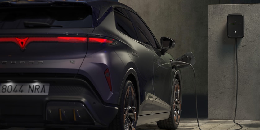 Rear view of a dark CUPRA Raval electric SUV plugged into a home wall charger in a concrete-walled garage