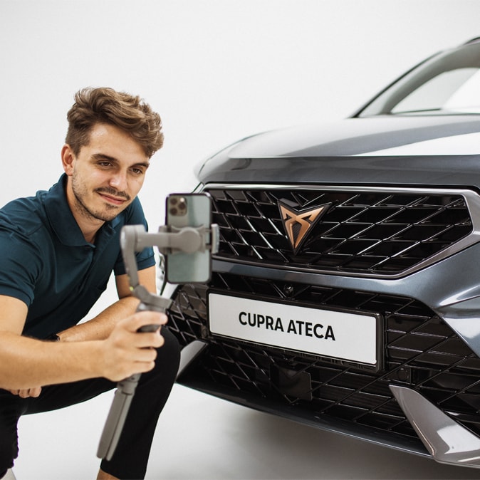 Osoba pozuje do zdjęcia z samochodem CUPRA Ateca, trzyma selfiestick, aby ująć w kadrze przód samochodu.