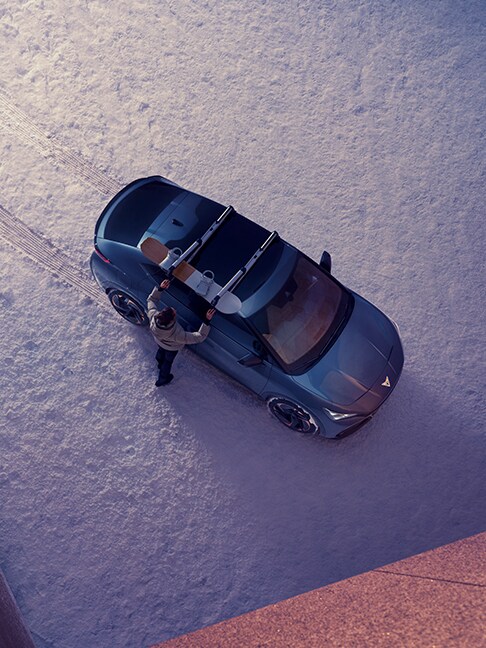 p-down view of a CUPRA Tavascan Fully electric SUV Coupé in Tavascan Blue, fitted with a roof ski rack in a snowy winter car park.