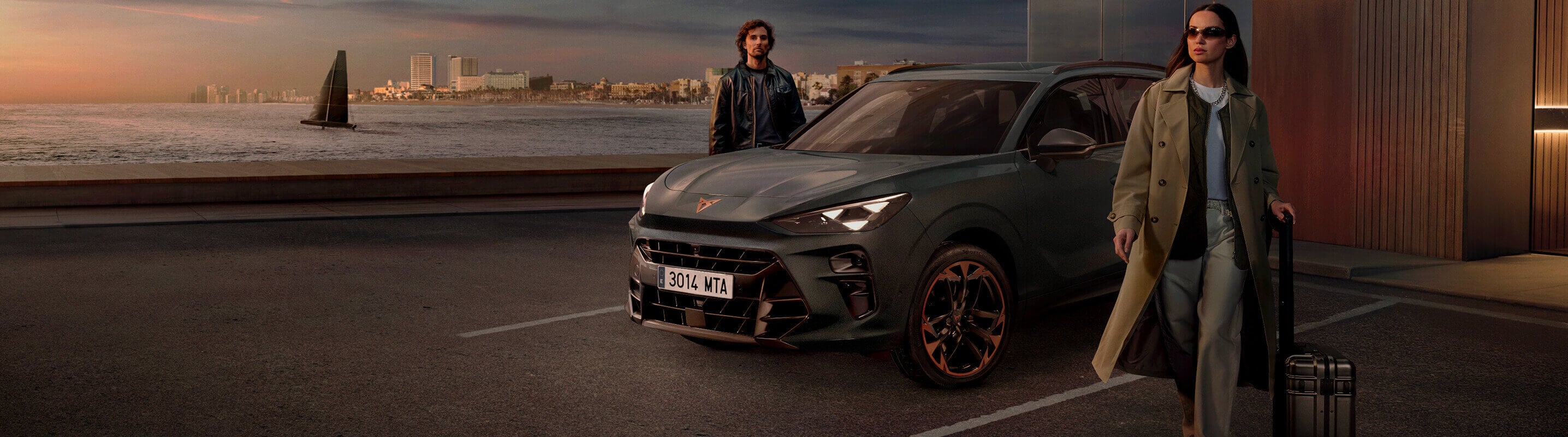 CUPRA Terramar SUV in Fiord Blue parked outside a hotel at night, with a man standing beside it and a coastal cityscape illuminated in the background.