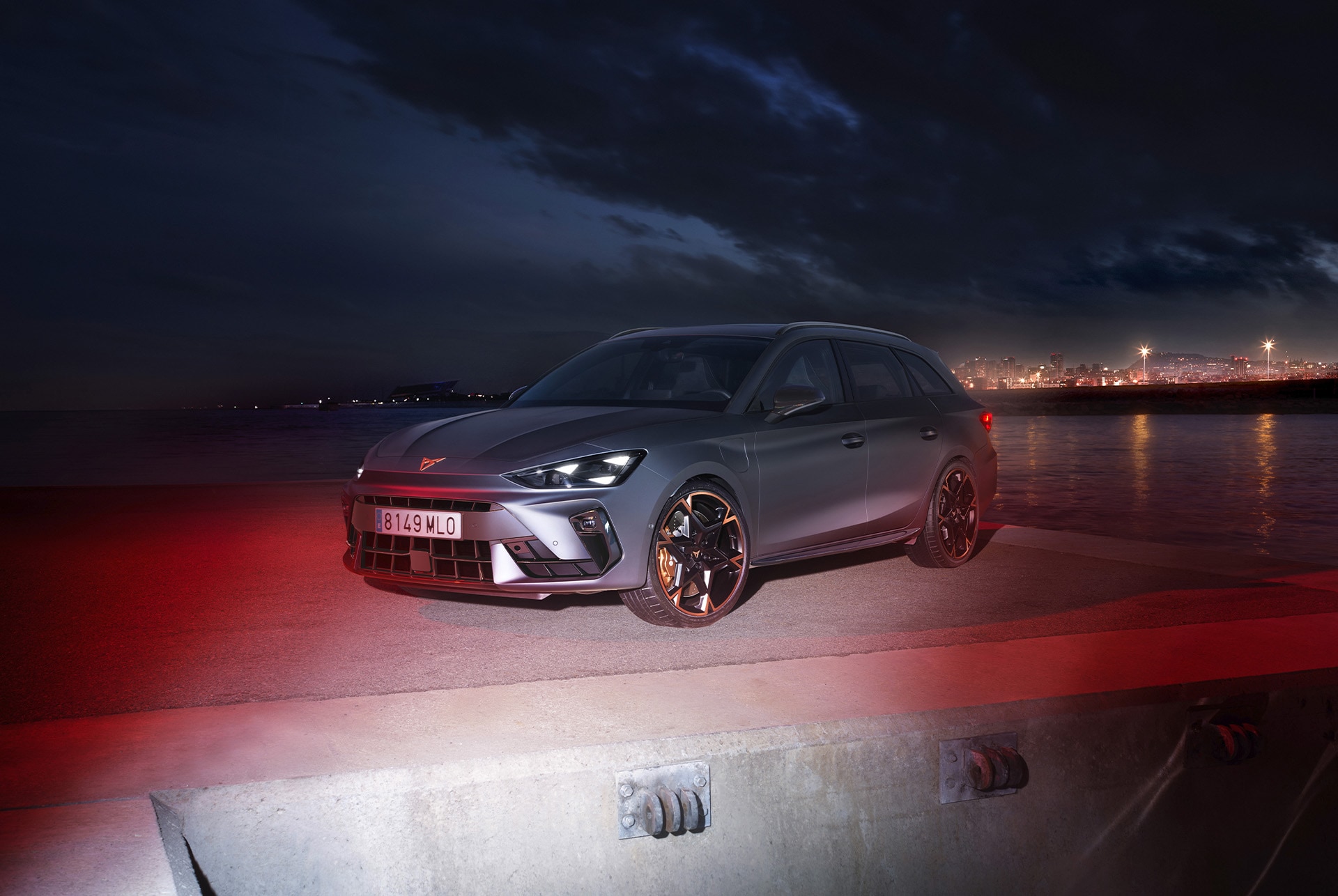 3/4 front view of the CUPRA Leon SP in Magnetic Tech Matt Grey with an urban seaside backdrop at night, showcasing 19-inch alloy wheels in Polar Copper.