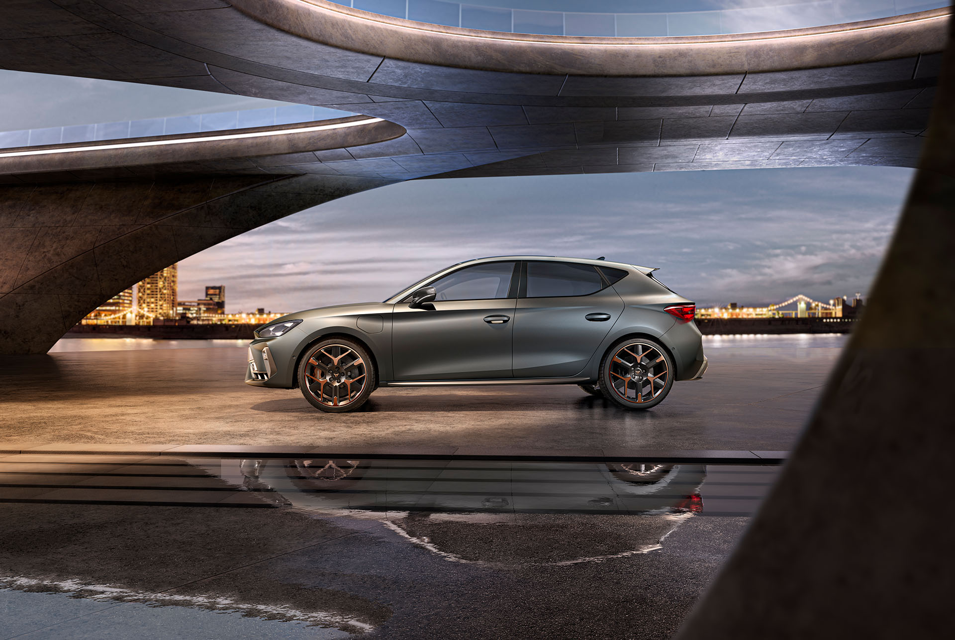Wide shot of new CUPRA Leon  side view in grey parked on smooth concrete under architectural structure with view of the sea and cityscape, cloudy sky.