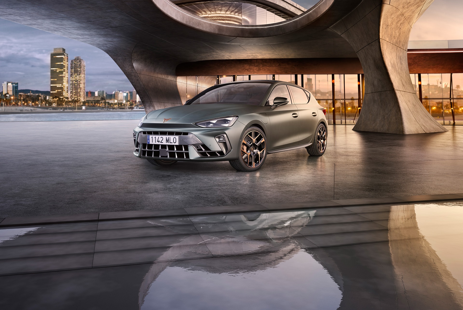 A new CUPRA Leon  in enceladus grey matte with copper accent alloy wheels parked on smooth concrete under an architectural structure. Barcelona cityscape in the background.