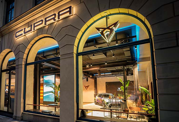 Beleuchtete Außenansicht der CUPRA City Garage München mit großem Logo und Blick auf den Showroom.