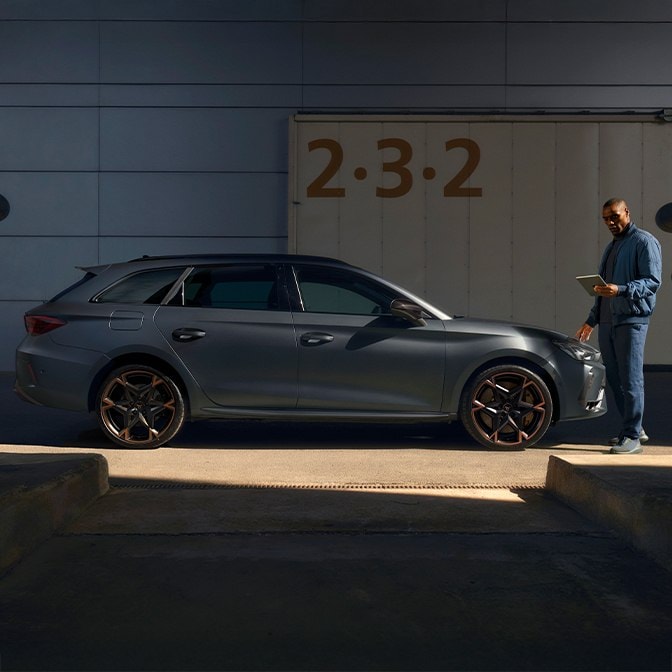 A side profile view of a matte grey CUPRA Leon featuring signature copper-accented alloy wheels, parked within a concrete industrial loading bay. A man in a blue jacket stands at the front of the vehicle, looking down at a digital tablet while sunlight casts sharp shadows across the scene.