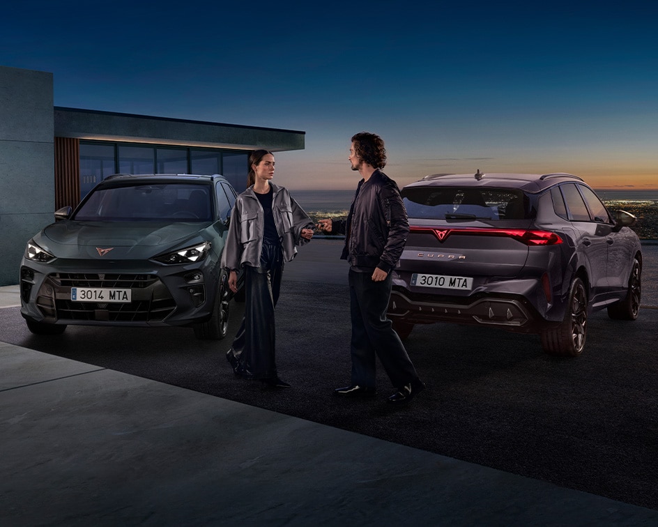 Two people standing between two parked CUPRA Terramar at twilight; a man on the right faces a woman on the left, who is standing near a dark green CUPRA Terramar and a wall-mounted charging station, while a purple Terramar is shown from the rear. 