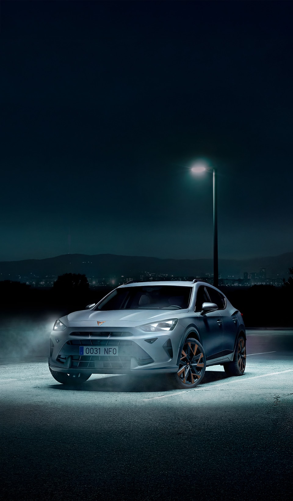 A front three-quarter view of a CUPRA Terramar parked in an empty outdoor parking lot at night. The vehicle is illuminated by a single overhead streetlamp, with mist or fog swirling around its front tires. The background features a dark silhouette of hills and distant city lights.