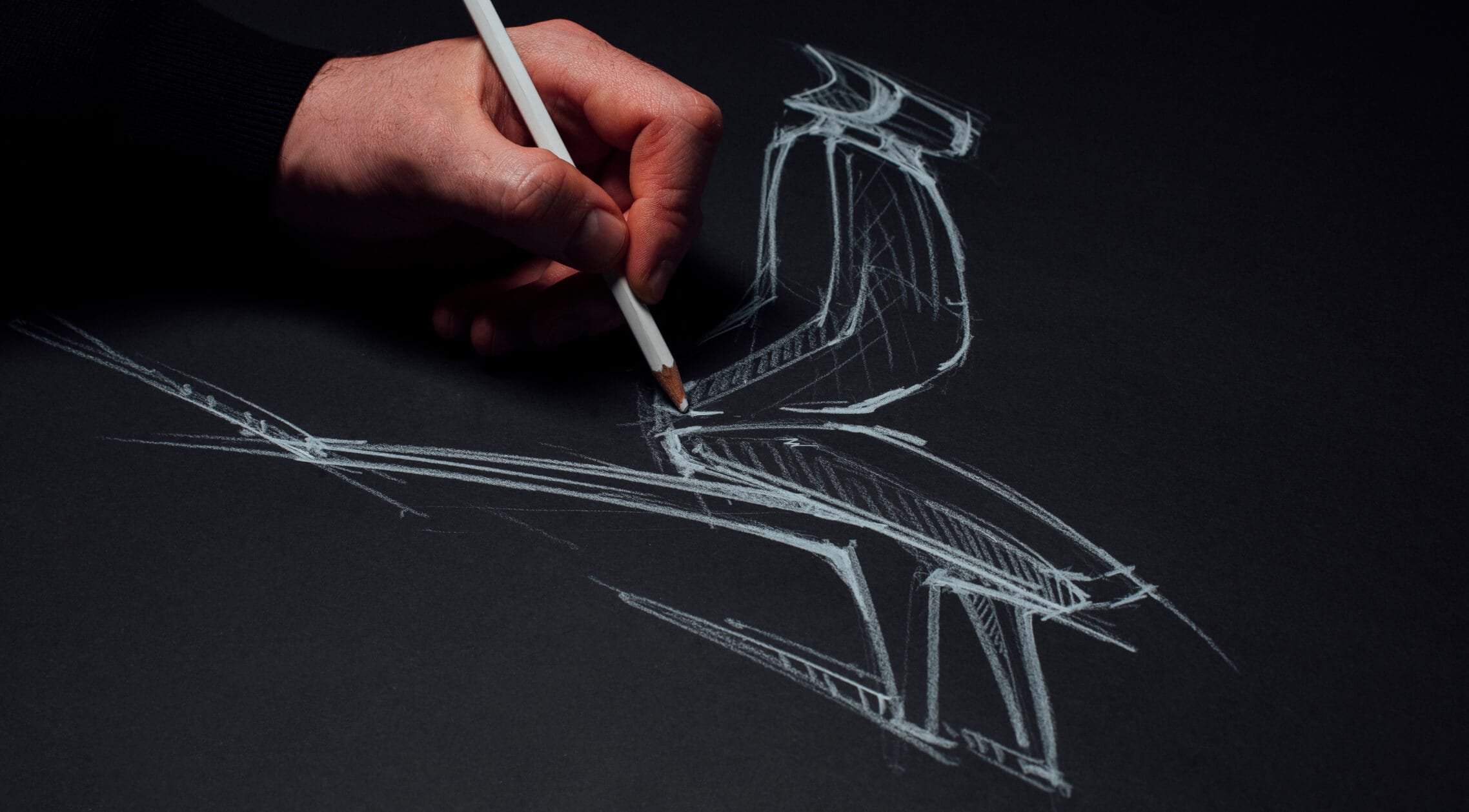 A hand holding a white pencil drawing a sketch of a chair with white lines on black paper.