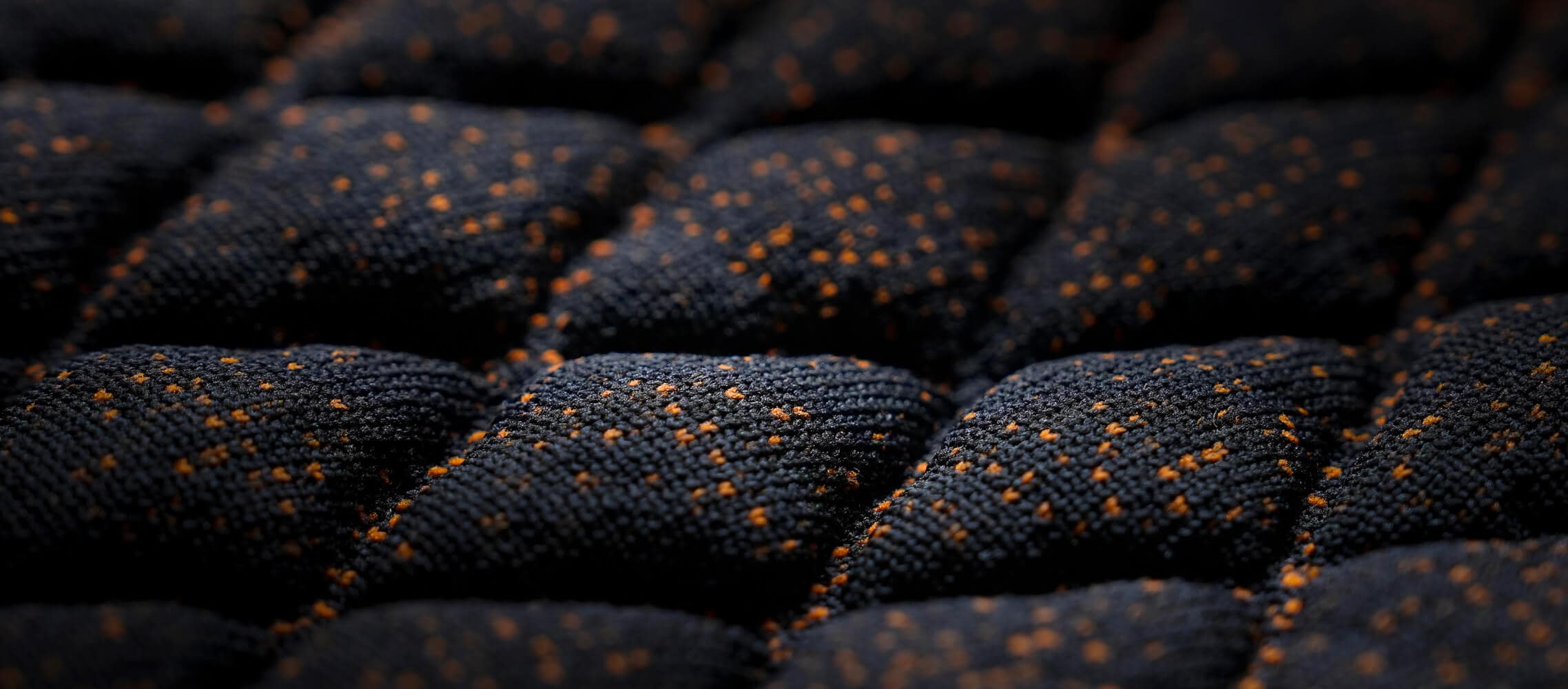 A close-up shot of a dark, quilted fabric with a raised diamond pattern and small orange flecks. 