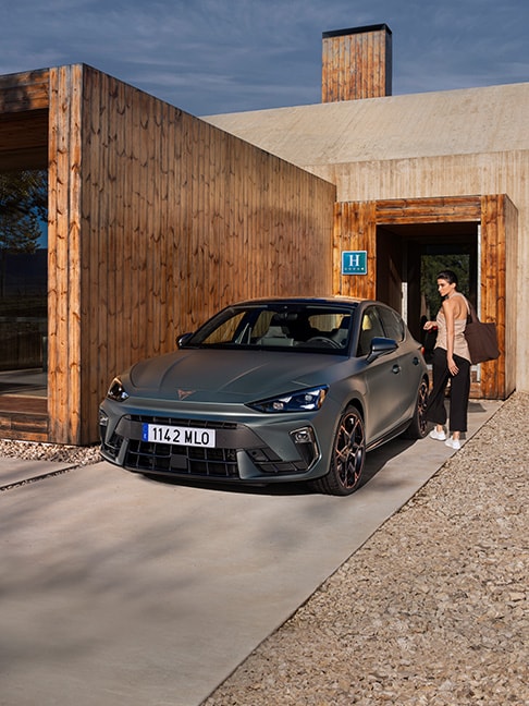 Un CUPRA León e-Hybrid compacto en color Gris Enceladus Mate frente a un hotel de lujo en verano.