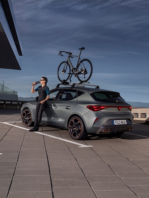 Un CUPRA León e-Hybrid compacto en Gris Enceladus Mate, con una bicicleta sobre las barras del techo, aparcado mientras un ciclista bebe agua de una botella.