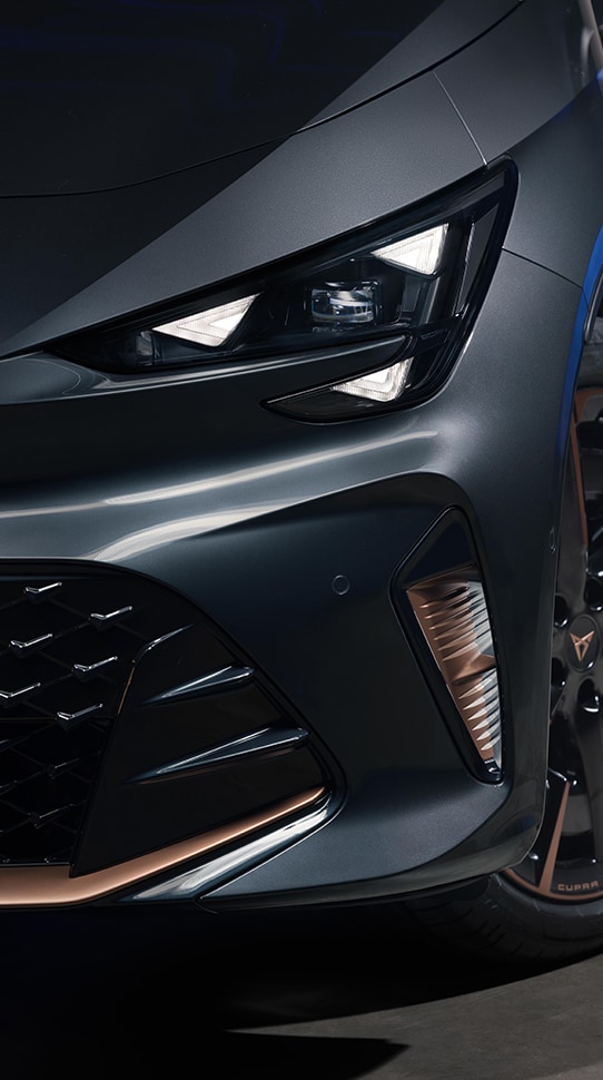 Szczegółowe zbliżenie przedniego lewego reflektora i zderzaka modelu CUPRA Born VZ w kolorze Timanfaya Grey,pokazujące trójkątne światła do jazdy dziennej LED oraz miedziane akcenty na dolnej kratce i felgach aluminiowych.