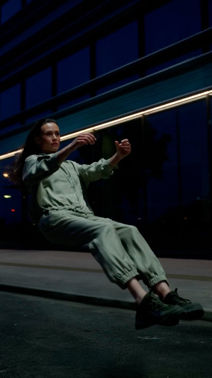 Une femme vêtue d'une combinaison vert clair et de baskets foncées semble flotter en position assise dans une rue de la ville la nuit, avec un immeuble moderne en verre en arrière-plan.
