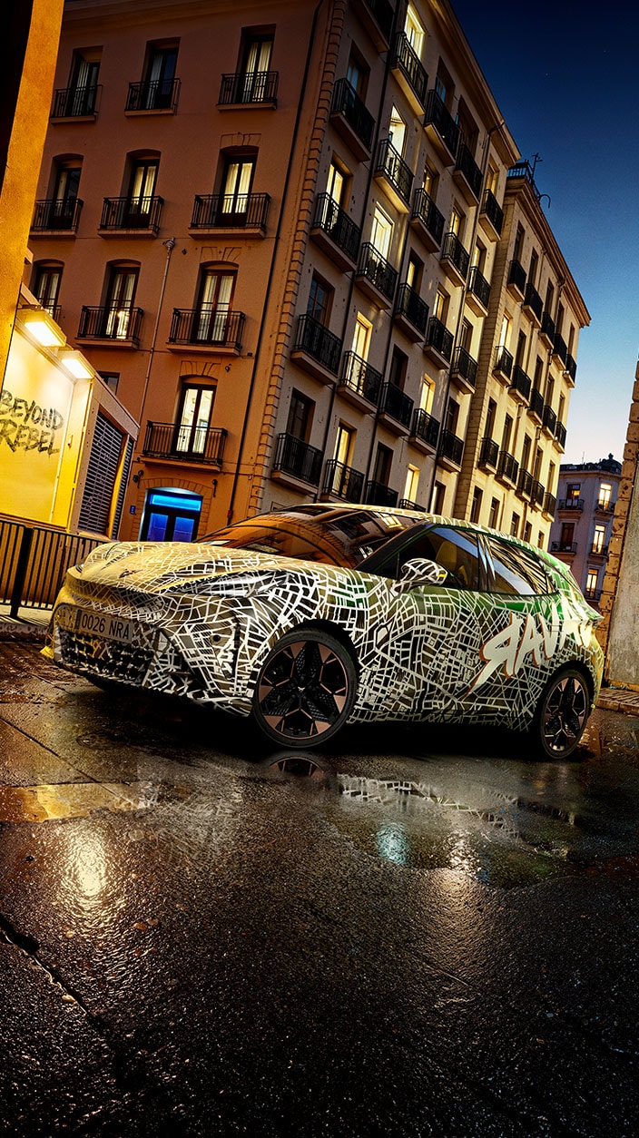 A CUPRA Raval camouflage wrap featuring abstract patterns and the word “Raval” parked on a wet street at dusk, with tall apartment buildings illuminated in the background.