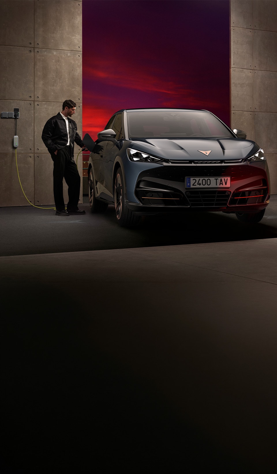 Front view of a CUPRA Raval parked in front of an illuminated modern building at night, with a woman sitting on the hood.
