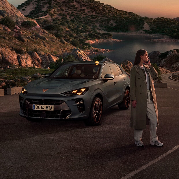 A CUPRA vehicle parked on a coastal road at sunset, with a person wearing a trench coat standing next to it, looking towards the sea.