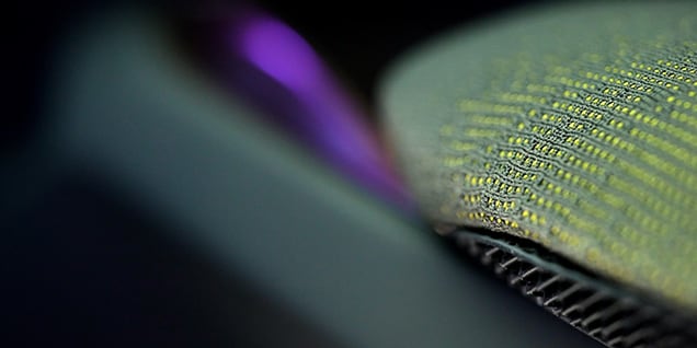 The image shows a macro (close-up) photograph of a textured surface. The focus is on a material with a woven or knitted pattern, where small yellow threads are interlaced with gray or greenish fibers.